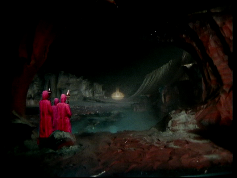 three robed figures in dark alien caves moving towards a sarcophagus