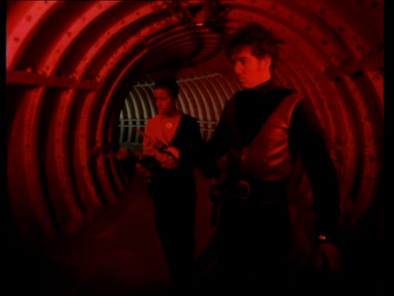 Dayna and Tarrant standing in a corridor with red lighting