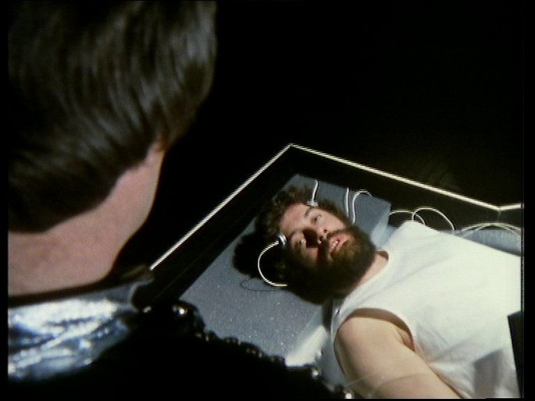 A bearded Blake lying down, hooked up to wires, staring up at Avon