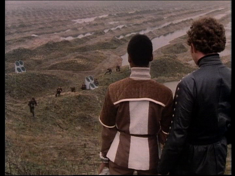 Dayna and Tarrant look down across the muddy hills