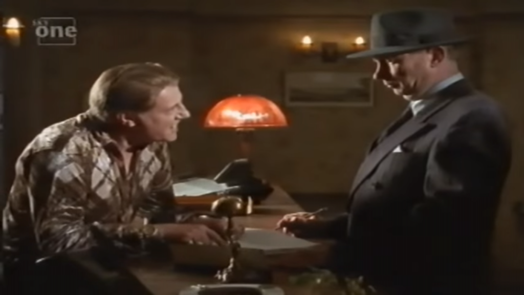 CD leaning over hotel reception desk to flynn