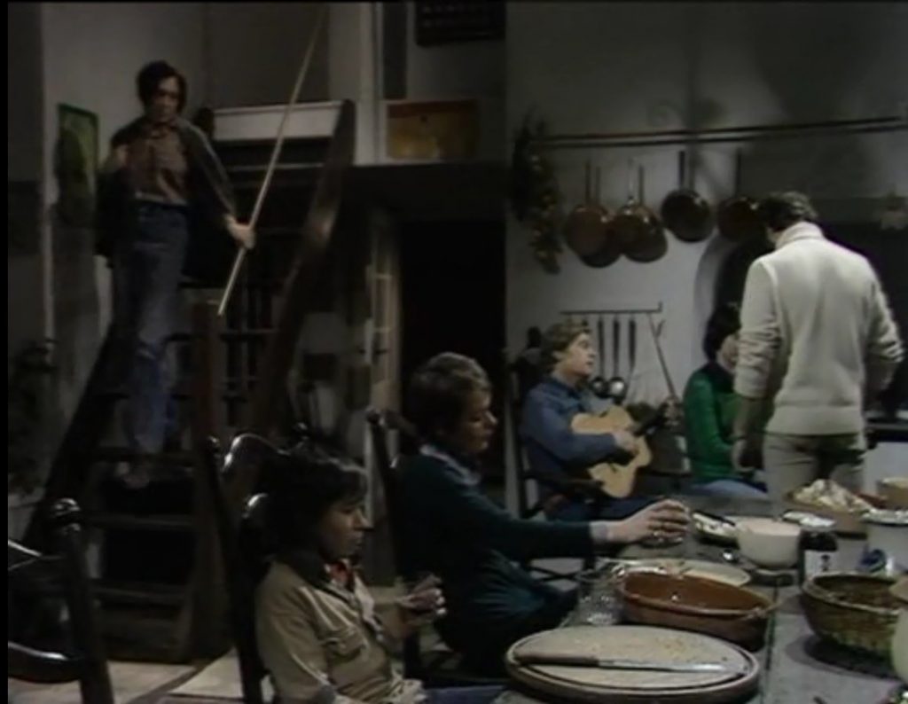 charles comes down the stairs while the others sit at the table drinking wine. greg is in the background playing guitar