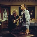 Doctor Schultz examines Callan's broken ribs as he sits on the surgery couch with a gun placed down beside him