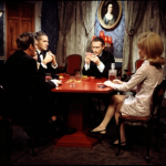 Miller, Lindale, Callan and Caroline sit around a hexagonal table playing cards. A whiskey decanter is next to Callan.