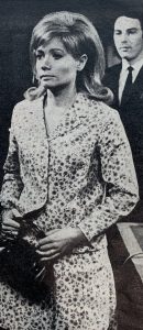 Anne stands in the foreground clutching her handbag with Meres over her shoulder in the background