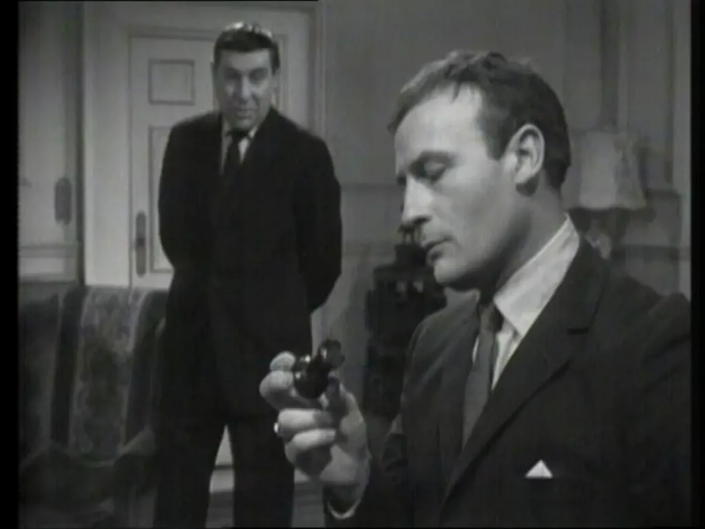 Bunin stands watching Callan as Callan thoughtfully plays with a chess piece